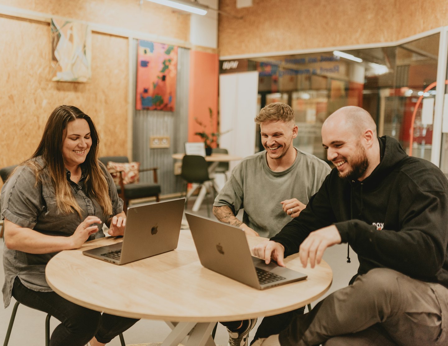 Three professionals collaborate on laptops around a wooden table.